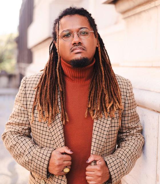 Jonas Davila leaning against a stone wall, holding his blazer lapel